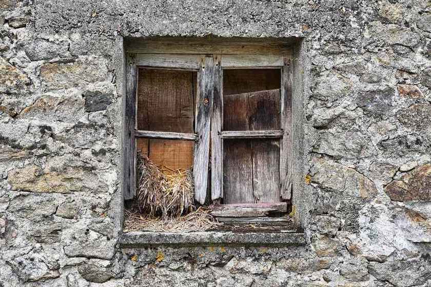 Fenêtre en bois décrépit enchâssée dans un mur en pierre grise et fissurée, avec du foin visible à l'intérieur Fenêtre en bois décrépit enchâssée dans un mur en pierre grise et fissurée, avec du foin visible à l'intérieur